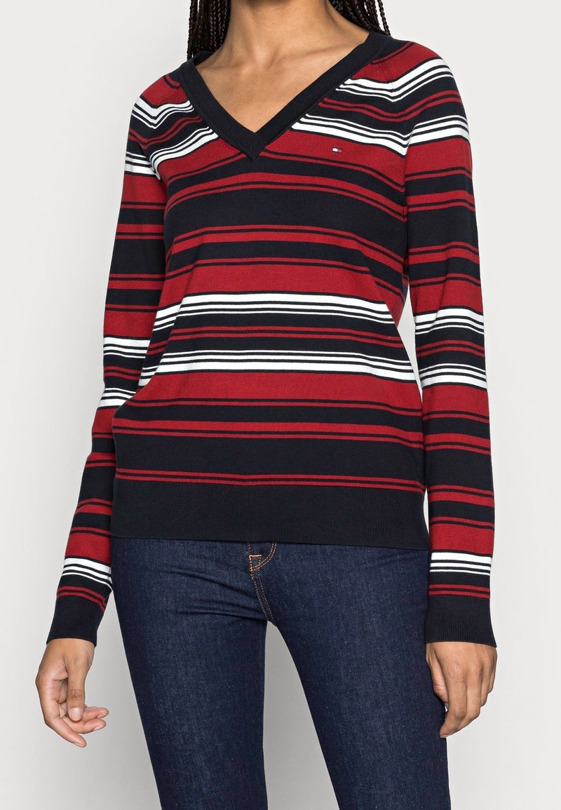 Femme portant un pull à rayures rouges, noires et blanches avec un col en V et des manches longues, associée à un jean bleu foncé, debout devant un fond clair uni.