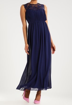 Robe maxi bleu marine avec un col en dentelle, un buste plissé et une jupe fluide. La mannequin l'a associée à des chaussures à talons hauts roses.