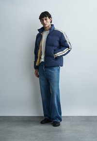 Junger Mann steht, trägt eine blaue gesteppte Jacke mit weißen Streifen, weißes Hemd, weite blaue Jeans und schwarze Schuhe vor einem einfarbigen Hintergrund.