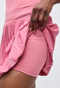 Χέρι σηκώνει ροζ πλισέ φούστα για να δείξει ροζ εφαρμοστά σορτς με πλαϊνή τσέπη και λογότυπο κοντά στο τελείωμα.