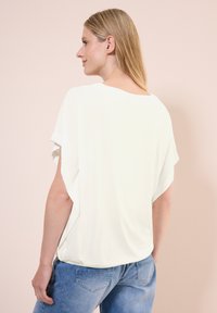 Bílý volný top s krátkými rukávy a kulatým výstřihem, vyrobený z měkké, hladké tkaniny. Nosený s světle modrými džíny.