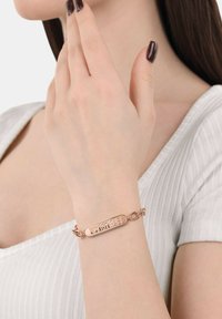 Pulsera en oro rosa con una placa rectangular grabada con la palabra "Escorpiones". Conectada por una cadena con eslabones redondos. Acabado liso.