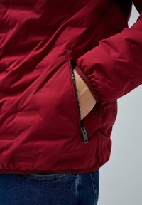 Mano dentro del bolsillo con cremallera abierto de una chaqueta acolchada roja, con jeans azules visibles abajo, fondo neutro.