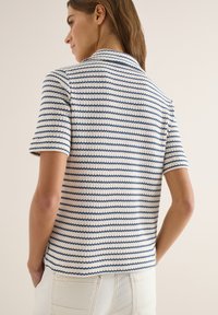 Femme portant un haut à manches courtes en maille blanche à rayures horizontales bleu marine, associé à un pantalon blanc taille haute, vue de dos.