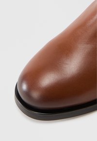 Chaussure en cuir marron avec une texture lisse, un bout rond et une semelle noire contrastante. Le design est élégant et minimaliste.