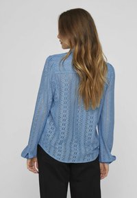 Blouse en dentelle bleu clair avec des motifs détaillés, des manches longues, un col boutonné et un ourlet arrondi. Portée avec un pantalon noir.