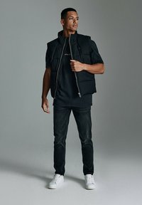 Czarna ocieplana kamizelka na czarnym t-shircie, zestawiona z ciemnymi dżinsami i białymi trampkami. Kamizelka ma wysoki kołnierz i zamek z przodu.