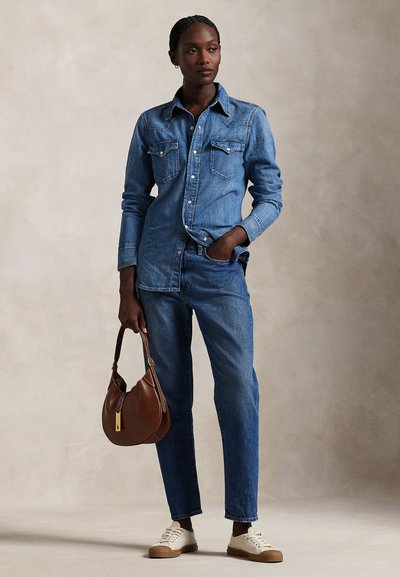 Femeie purtând o cămașă din denim cu nasturi și blugi, ținând o poșetă maro în mâna stângă, mâna dreaptă în buzunar, stând pe un fundal simplu.