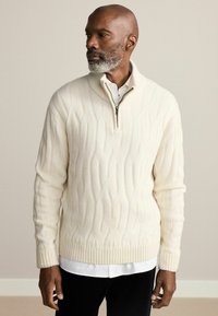 Cream-colored, cable-knit sweater with a quarter-zip collar, ribbed cuffs, and hem. Worn over a white button-up shirt.