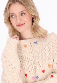 Maglione a maglia color crema con motivi a cuore in vari colori, caratterizzato da un motivo testurizzato e polsini a coste.