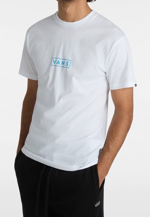 Mężczyzna w białej koszulce Vans z niebieskim logo na piersi i czarnych spodniach Vans, ręce w kieszeniach, na jednolitym jasnym tle.