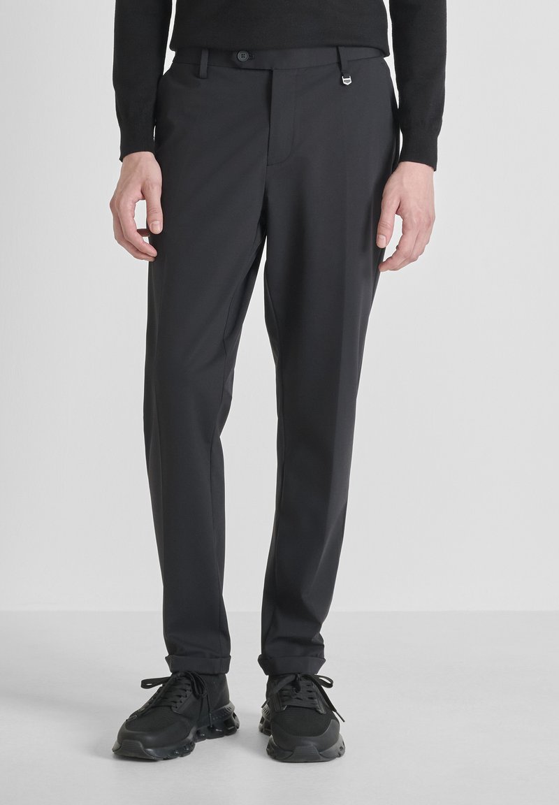 Uomo che indossa pantaloni neri attillati e sneakers nere in piedi contro uno sfondo chiaro e semplice.