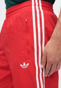 Rode atletische shorts met witte strepen aan de zijkanten, voorzien van een zak met rits en een geborduurd logo op het onderbeen. Lichtgewicht materiaal.