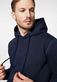 Ciemnoniebieska bluza z kapturem, wykonana z miękkiego materiału, z przednią kieszenią typu kangur, regulowanymi sznurkami i haftowanym logo na piersi.