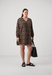 Μαύρο και μπεζ φλοράλ φόρεμα με μακριά μανίκια και V λαιμόκοψη, φορεμένο με μαύρες σαγιονάρες. Κρατά μια μαύρη τσάντα tote.