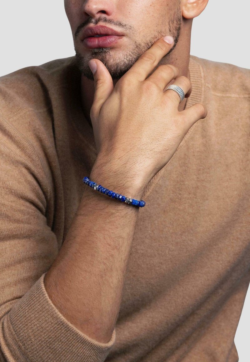 Blue beaded bracelet with varied shapes, featuring smooth and textured stones, worn on a wrist, paired with a beige sweater.