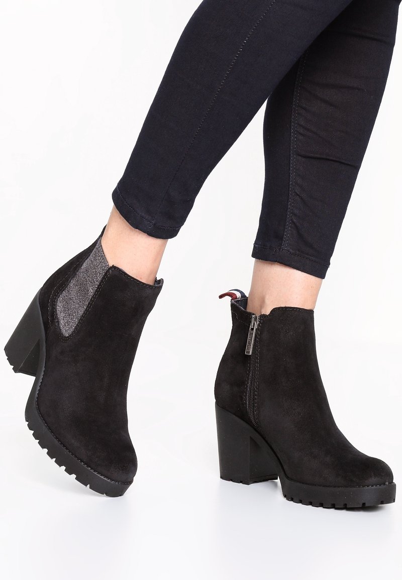 Personne portant des bottines en daim noir à talons épais et fermeture éclair latérale, associées à un jean skinny foncé.