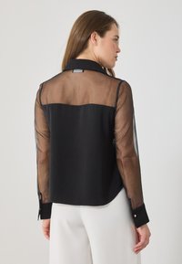 Μαύρη μπλούζα με διαφανείς μακρύ μανίκια, συμπαγές πίσω μέρος και σχέδιο με γιακά. Διαθέτει κουμπωτές μανσέτες και λεία υφή.