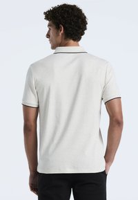 Polo-shirt à manches courtes en beige avec une finition texturée, présentant une bordure noire au niveau du col et des ourlets des manches. Design épuré avec une coupe droite.