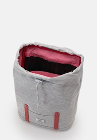 Grå tygdragsäck med röda detaljer inuti. Har ett randigt fodral och plastdetaljer. Slät yttextur.