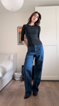 Junge Frau in einem schwarzen Langarmshirt und weitem blauen Jeans, die in einem Schlafzimmer mit Bett und gerahmtem Poster im Hintergrund steht.