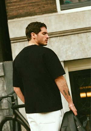 Mann mit kurzen dunklen Haaren und Tattoos am linken Arm trägt schwarzes Hemd und weiße Hose, hält schwarze Jacke und steht bei einem Fahrrad neben einem Gebäude.