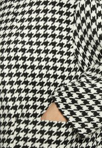 Czarna i biała tkanina w wzór houndstooth, charakteryzująca się miękką teksturą i boczną kieszenią. Wzór jest wyrazisty i dobrze kontrastujący.