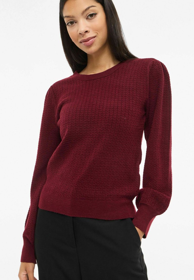 Bordowy sweter dziergany o fakturowanej powierzchni, z okrągłym dekoltem i długimi rękawami. Posiada dopasowane mankiety i prosty dół.