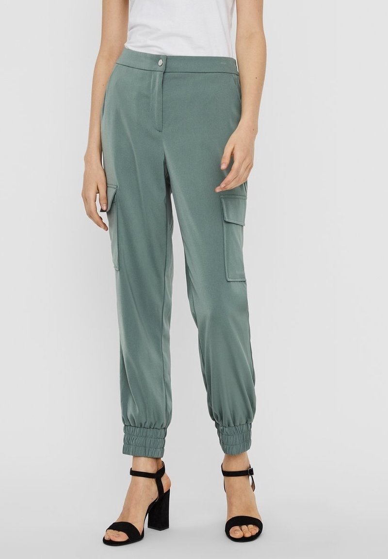 Femme portant un pantalon cargo vert avec des poignets élastiques et des sandales noires à talons hauts, assortis à un haut sans manches blanc.
