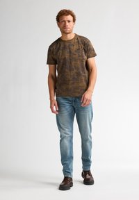 Bruine camouflagepatroon korte mouwen shirt, lichtblauwe spijkerbroek en bruin veterschoenen. Glad stof, losse pasvorm, rechtopstaand.
