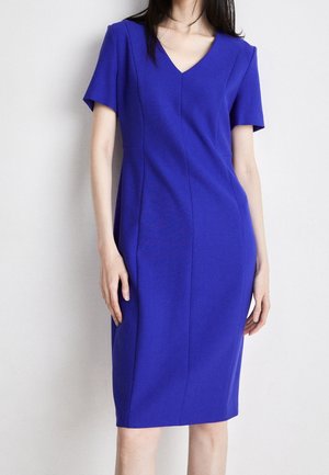 Woman wearing a fitted, knee-length, short-sleeve royal blue dress with a V-neckline, standing against a plain light background.