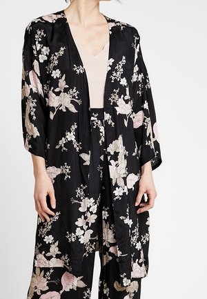 Femme portant une veste style kimono noire fleurie et un pantalon assorti sur un haut beige clair, montrée du cou jusqu'à mi-cuisse.