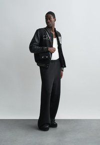 Schwarze Leder-Collegejacke mit weißer Stickerei, kombiniert mit lockeren schwarzen Hosen. Das Modell steht vor einem hellgrauen Hintergrund.