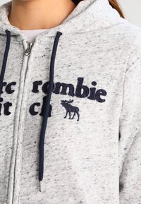 Felpa grigia con zip realizzata in tessuto morbido, caratterizzata da scritte in navy e un logo di alce blu. Include stringhe con punte in metallo.