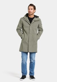 Un manteau imperméable vert olive clair avec capuche, fermeture éclair à l'avant et poches latérales, associé à un jean bleu et des baskets noires.