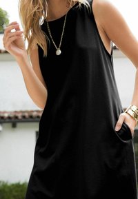 Robe noire sans manches au coupe décontractée, dotée de poches latérales. Le mannequin porte un collier en chaîne dorée avec un pendentif rond.