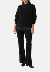 Kobieta ubrana w czarny dzianinowy sweter na czarnej koszuli zapinanej na guziki, do tego czarne szerokie spodnie i czarne botki na obcasie.