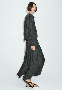 Robe longue gris foncé au design fluide, dotée de longues manches et d'une coupe ample. Portée avec des tongs noires, mettant en valeur son style décontracté.