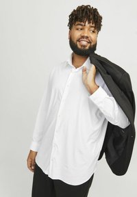 Weißes Langarm-Hemd mit Knöpfen in lockerer Passform, kombiniert mit einem schwarzen Blazer über der Schulter. Der Hintergrund ist neutral grau.