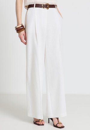 Femme portant un pantalon blanc taille haute à jambes larges avec une ceinture marron, des sandales à talons marron et des bracelets marron empilés au poignet gauche.