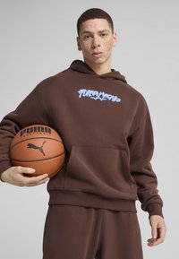 Hanorac maro cu buzunar frontal, având logo-ul albastru "Puma Hoops". Modelul ține o minge de baschet, îmbrăcat în pantaloni de trening maro asortati.