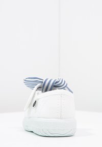 Zapatilla de lona blanca con forma redondeada, suela de goma texturizada y un lazo de tela a rayas azul y blanco en la parte trasera.