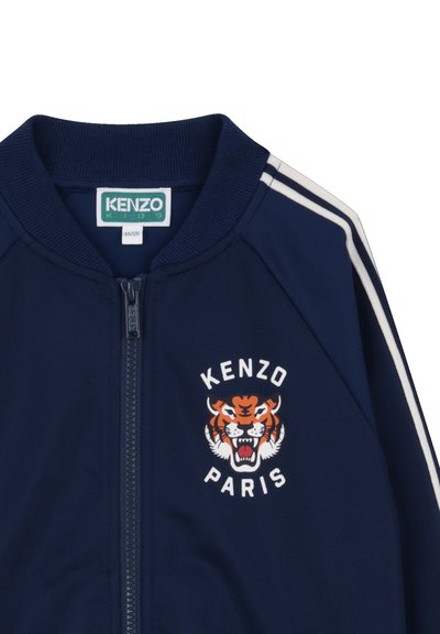 Marineblauwe ritssluiting jas met geribde kraag, voorzien van een tijgerafbeelding en "KENZO PARIS" in het wit gedrukt. Witte strepen op de mouwen.