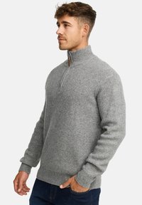 Mand iført en grå strikket sweater med lynlås og høj krave samt mørkeblå jeans, står med den ene hånd i lommen og ser til venstre.