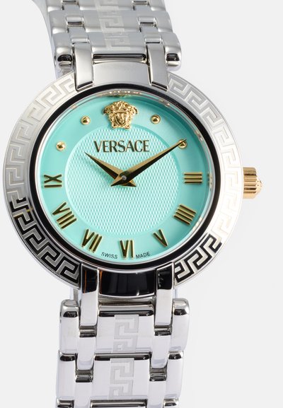 Versace pulkstenis ar apaļu sudraba korpusu, piparmētru zaļo teksturēto ciparnīcu, zeltainiem akcentiem un rakstainu sudraba aproci. Melnie grieķu atslēgu elementi gar apakšmalu.