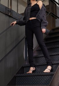 Chaqueta de mezclilla negra corta sobre una camiseta sin mangas negra, combinada con jeans ajustados negros y sandalias de tacón alto negras con tiras. Escalera metálica empinada.