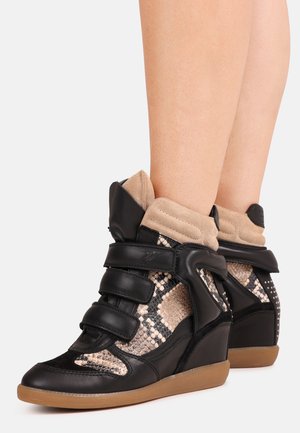 Zapatillas de cuña negras y beige con paneles con estampado de serpiente y tres tiras de velcro, usadas en piernas desnudas contra un fondo liso.