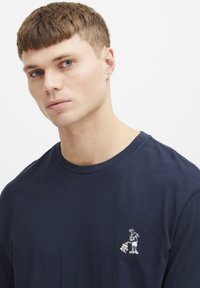 Marineblå bomullst-skjorte med et lite hvitt og gult broderi på venstre bryst. Klassisk rund hals og korte ermer.