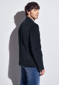 Black corduroy blazer with a tailored fit, featuring a single vent and buttoned cuffs, paired with blue denim jeans.