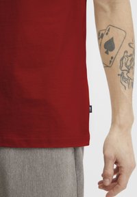 Red cotton t-shirt features a straight hem and side tag; a textured gray trouser fabric visible below with a skin-covered, tattooed arm.